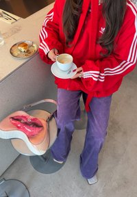 Άτομο με κόκκινο σακάκι Adidas και μωβ παντελόνι κρατάει ένα φλιτζάνι καφέ και πιατάκι, καθισμένο δίπλα σε ένα τραπέζι με ένα κρουασάν και μια τσάντα με στάμπα από χείλη.