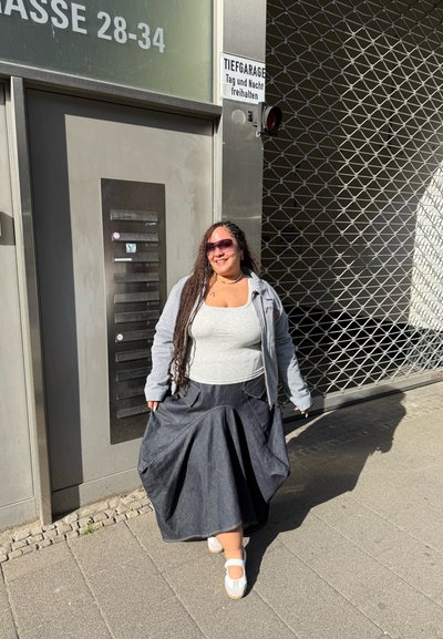 Mujer con el cabello largo trenzado que lleva gafas de sol, chaqueta gris, camiseta blanca y falda oscura, de pie sonriendo en la acera cerca de una puerta de garaje cerrada.