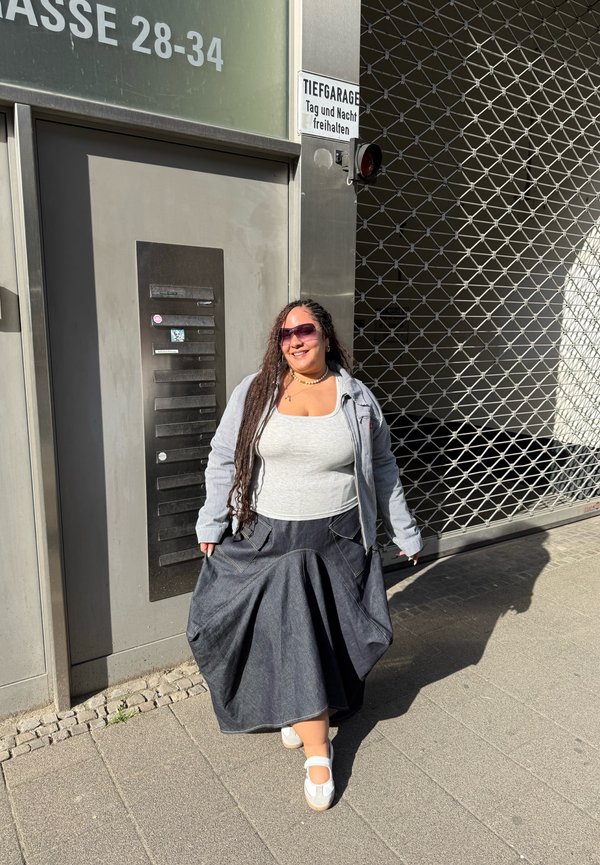 Žena s dlhými zapletenými vlasmi, ktorá má na sebe slnečné okuliare, sivú bundu, biely top a tmavú sukňu, stojí usmiata na chodníku blízko zatvorenej garážovej brány.