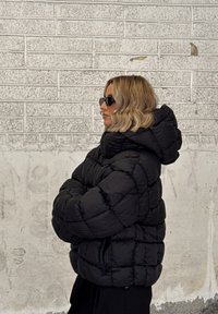 Fekete puffamellény steppelt mintával és kapucnival, fényes textúrával. Fekete napszemüveggel viselve, semleges háttér előtt.