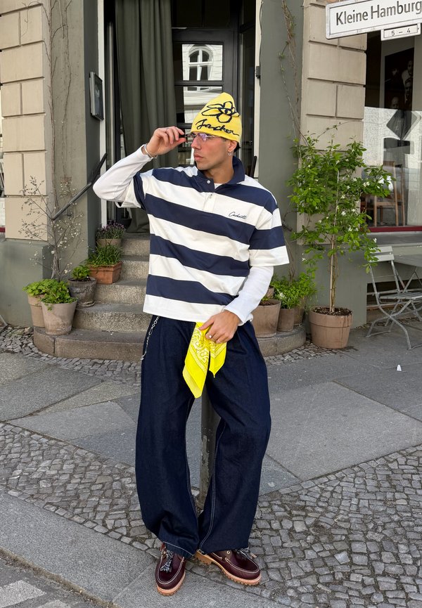 Mann med gul lue og stripete skjorte holder et gult bandana, står på en brosteinsbelagt fortau nær en bygninginngang med potteplanter.