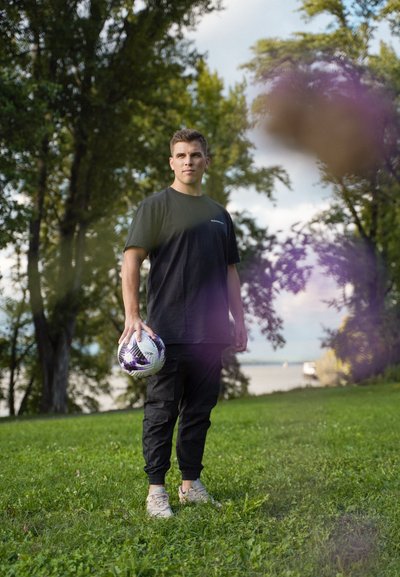 Camiseta negra y pantalones cargo negros llevados por una persona que sostiene un balón de fútbol con un diseño. Césped y árboles de fondo, cielo nublado arriba.