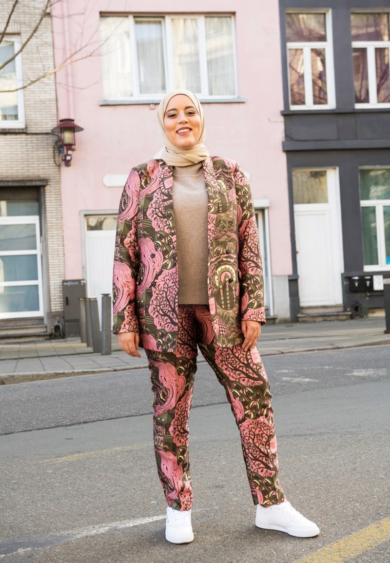 Patterned suit in green and pink with floral designs, paired with a beige sweater and white sneakers. Model wearing a light headscarf.