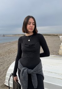 Jeune femme en robe noire se tenant sur une plage nuageuse, avec un pull gris noué autour de la taille et tenant un sac noir.