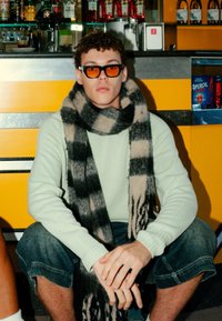 Person wearing oversized black and orange sunglasses, a light green sweater, and a black-and-beige checked scarf, seated with denim shorts.