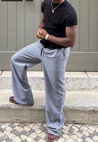 Bărbat îmbrăcat cu un tricou negru, o bijuterie de aur la gât, un ceas argintiu și pantaloni cu carouri largi, purtând pantofi maronii, stând cu un picior ridicat pe o treaptă de piatră.