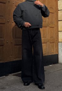 Personne portant une veste boutonnée gris foncé, un large pantalon noir et des chaussures noires, debout devant des portes en bois.