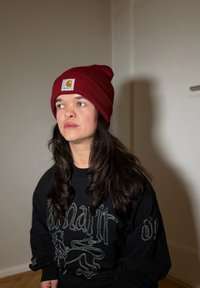 Czerwona dzianinowa czapka z metką z logo, zestawiona z czarną bluzą z białym haftem przedstawiającym stworzenie podobne do lwa oraz tekstem.