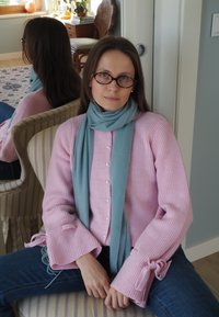 Svetloružový rebrovaný sveter s gombíkmi a dlhými rukávmi zdobenými mašličkami. Okolo krku je zaviazaný jemný modrý šál, k tomu riflové nohavice.