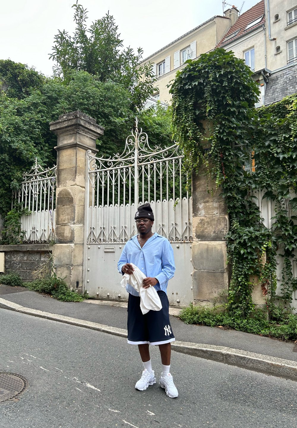 Ανοιχτό μπλε πουκάμισο με κουμπιά, μαύρες βερμούδες με λευκό λογότυπο, λευκά αθλητικά παπούτσια. Στέκεται μπροστά από μια σιδερένια πύλη, με πράσινη βλάστηση ορατή.