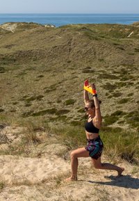 Entusiasta do fitness em top desportivo preto e calções estampados, fazendo um lunge em terreno arenoso com colinas gramadas e o oceano ao fundo. Banda de resistência brilhante acima da cabeça.