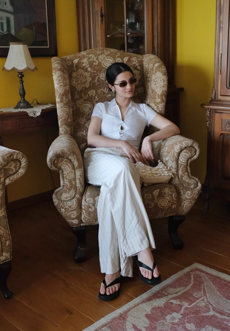 Femme portant des lunettes de soleil, un chemisier blanc et un pantalon rayé, assise détendue dans un fauteuil à motifs tenant un sac tressé dans une pièce style vintage.