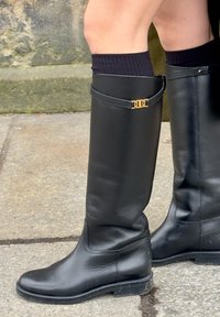 Femme portant des bottes en cuir noir montantes jusqu'aux genoux avec un emblème doré, associées à des chaussettes noires, debout sur un pavement en pierre près d'un mur de pierre.