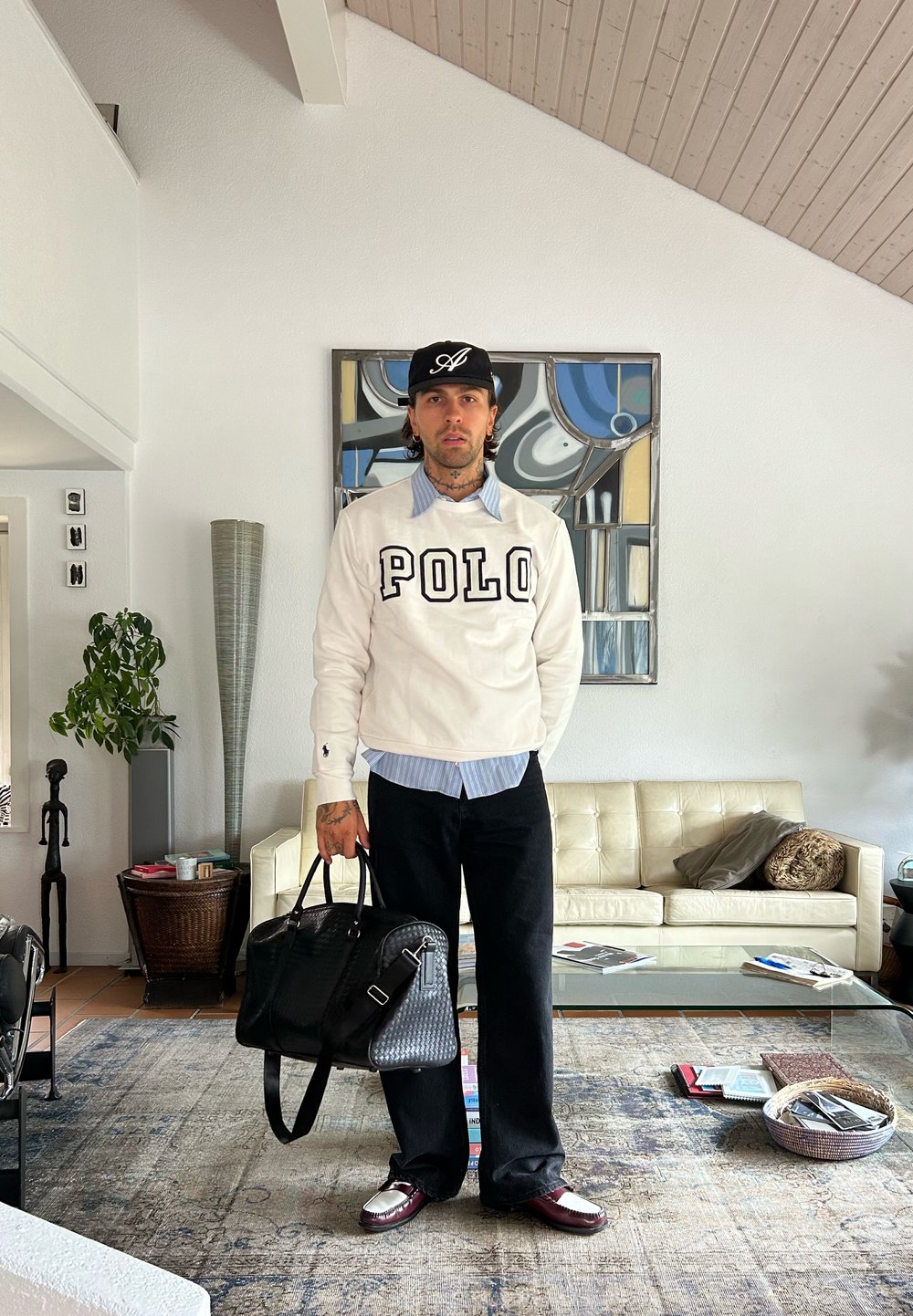 Mann steht im Wohnzimmer, trägt einen weißen "POLO"-Sweater, schwarze Hose und eine Kappe, hält eine große schwarze Tasche neben dem Sofa und dem Glastisch.
