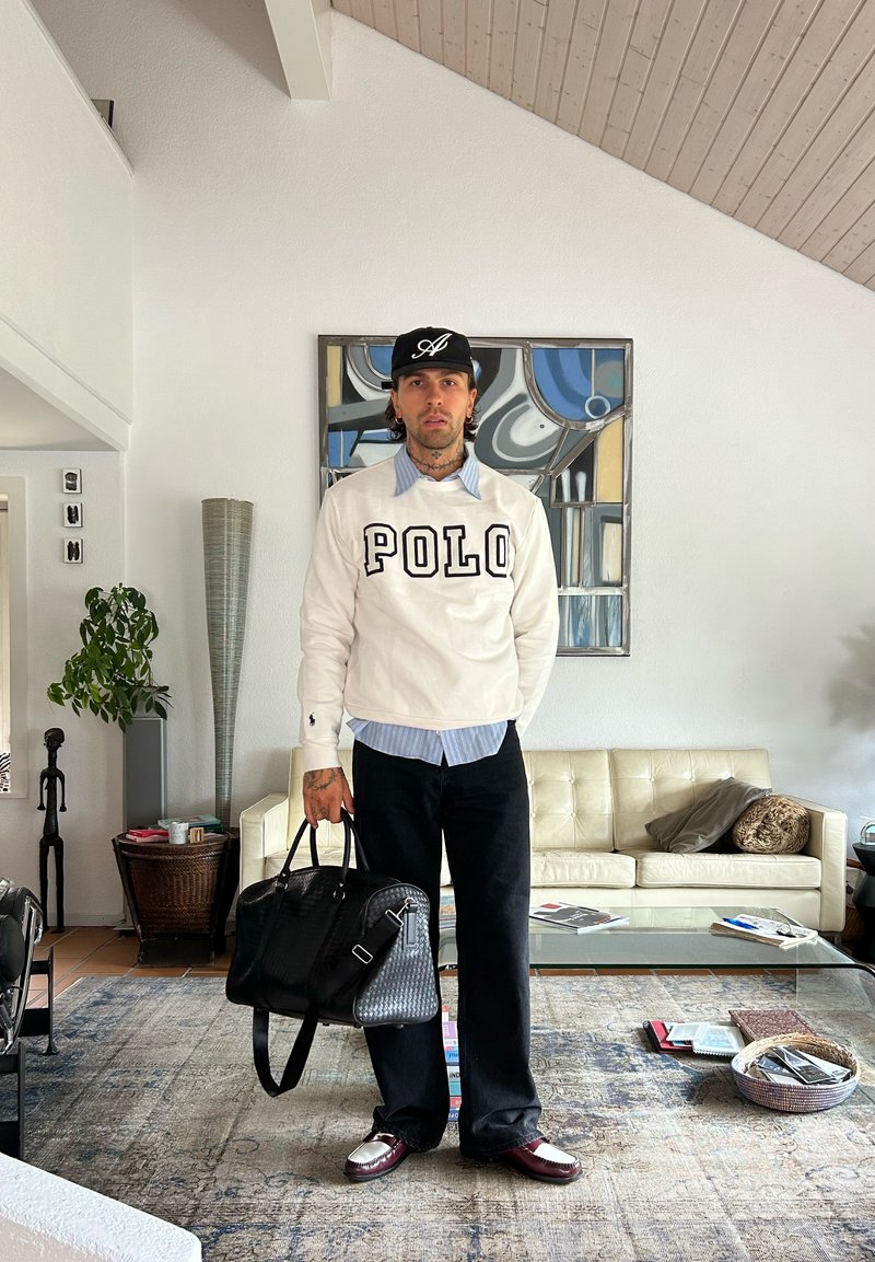 Homem está de pé na sala de estar, vestindo um sweatshirt branco "POLO", calças pretas e um boné, segurando uma grande bolsa preta ao lado do sofá e da mesa de café de vidro.