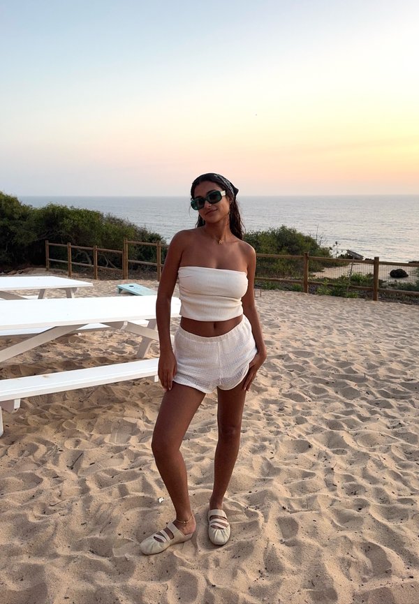 Mujer con top de tubo blanco y pantalones cortos, que lleva gafas de sol y pañuelo en la cabeza, está de pie en la playa de arena cerca de mesas de picnic al atardecer junto al mar.