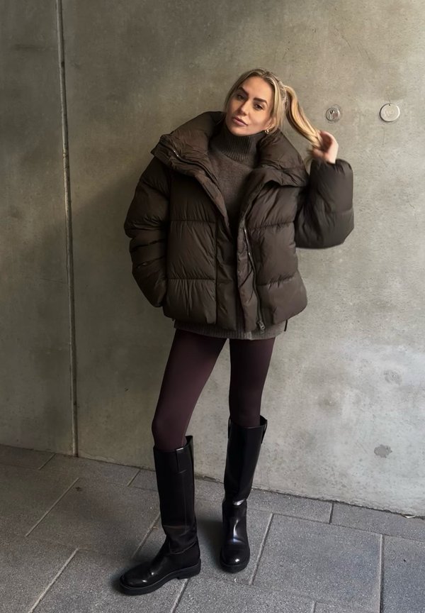 Bruine donsjas met een hoge kraag, gecombineerd met een donkerbruine coltrui, bordeauxrode legging en zwarte laarzen tot aan de knie. Gestructureerde betonnen achtergrond.