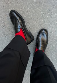 Schwarze Lederloafers mit strukturiertem Finish, getragen mit knallroten Socken, vor einem grauen Asphalt-Hintergrund. Schwarze Hosen sind teilweise sichtbar.