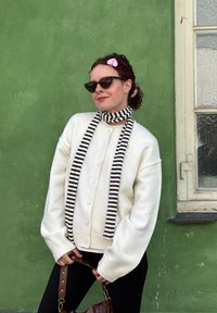 Liemenė, balta megzta su dideliais mygtukais, juodai baltas dryžuotas šalikas ir tamsūs saulės akiniai. Rudas krepšys su auksine furnitūra. Žalia fono spalva.