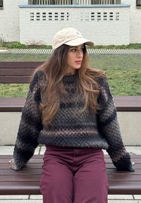 Νεαρή γυναίκα με μπεζ καπέλο και σκούνη πουλόβερ με σχέδια κάθεται σε παγκάκι εξωτερικού χώρου και κοιτάζει προς τα δεξιά της.