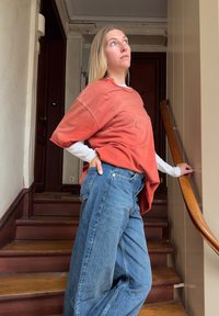 Νεαρή γυναίκα με μακριά ξανθά μαλλιά, φορώντας μεγάλη πορτοκαλί πουκάμισο και μπλε τζιν, στέκεται σε ξύλινη εσωτερική σκάλα, κοιτάζοντας προς τα πάνω, με το χέρι στο κιγκλίδωμα.