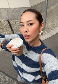 Raita villane cardigan tumesinine ja hallis, ringikujulised nööbid, hoiab papist kohvitassi, nähtav nahkrihmaga käekott.