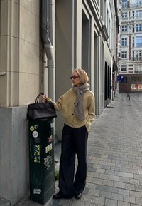 Béžový žebrovaný kardigan, šedý šátek, černá kabelka a široké pruhované kalhoty. Městské prostředí se zelenou utility skříní pokrytou nálepkami.