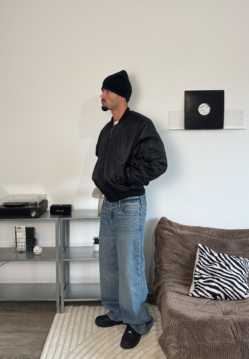 Sort bomberjakke med lynlåslukning, løstsiddende blå jeans, sort hue og sorte sko. Tekstureret brun sofa og pude med zebra-mønster i baggrunden.