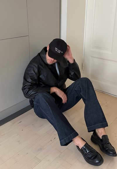 Hombre sentado en un suelo de madera clara contra la pared y una puerta cerrada, vistiendo una chaqueta de cuero negra, jeans oscuros, mocasines negros y una gorra negra.