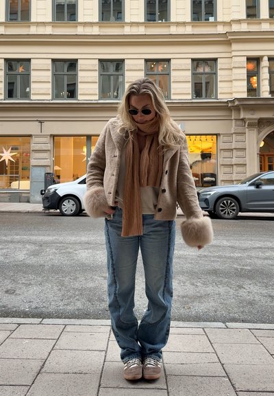 Mujer con chaqueta de piel beige con grandes puños, bufanda marrón, gafas de sol, vaqueros azules y zapatillas de deporte, de pie en la acera de la ciudad con edificios y coches al fondo.