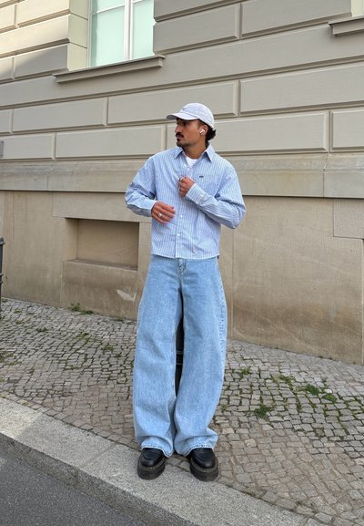 Camisa de botones a rayas azul y blanca, pantalones vaqueros oversized de color azul claro, y zapatos de plataforma negros. La figura lleva una gorra clara y auriculares.