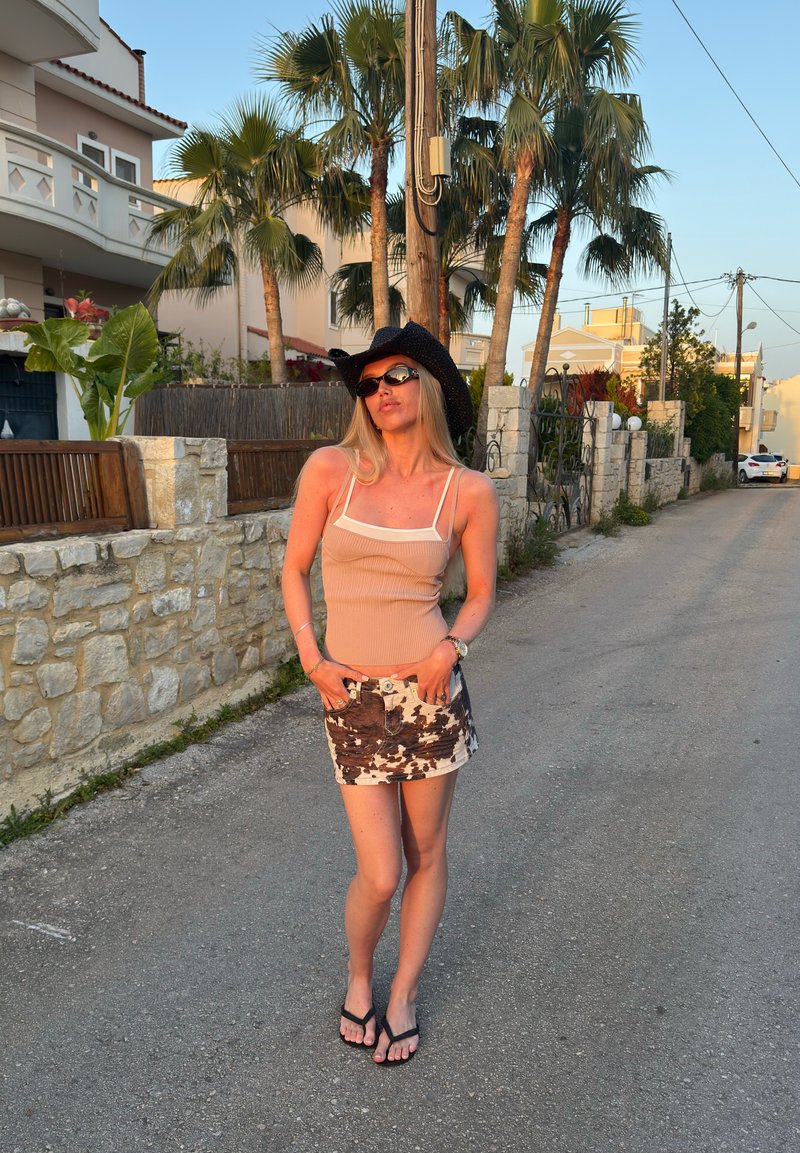 Jeune femme portant un chapeau de cowboy noir, des lunettes de soleil, un débardeur beige, une jupe à imprimé vache et des tongs, se tient dans une rue ensoleillée avec des palmiers.