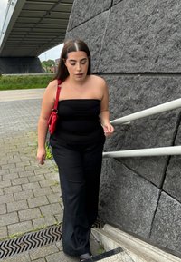 Svart bandeau-top, svarta vida byxor och svarta sandaler. Röd väska över axeln. Ljust grå stenvägg och trappor i bakgrunden.