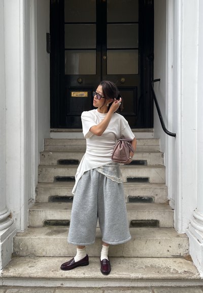 Mujer con top blanco y pantalones anchos grises de pie en escalones de piedra frente a una puerta negra, ajustándose el cabello con una mano.