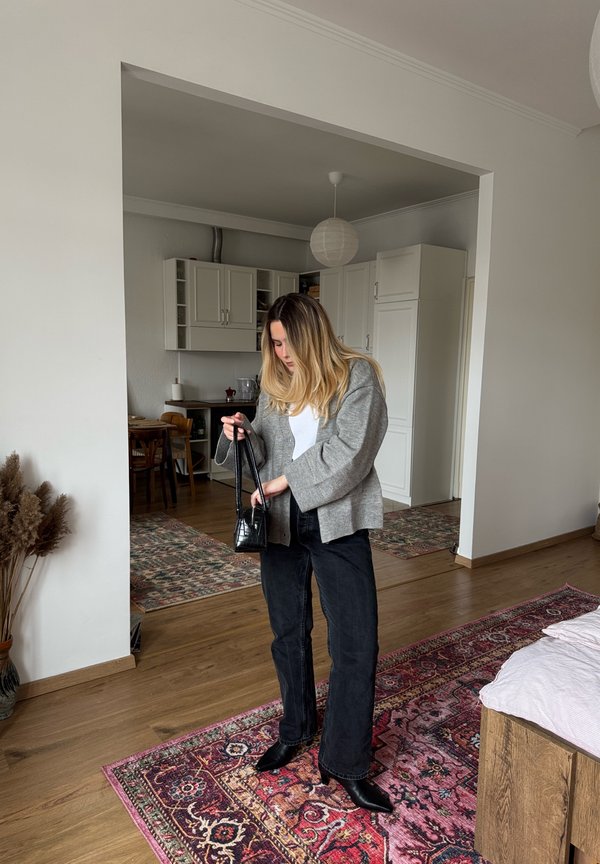 Žena v neformálním oblečení drží malou černou kabelku v moderním, uklizeném obytném prostoru s dřevěnými podlahami a vzorovanými kobercemi.