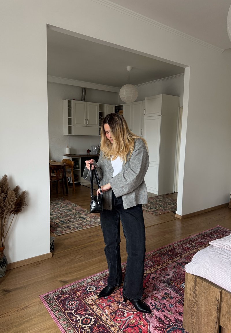 Femme en tenue décontractée tenant un petit sac à main noir dans un espace de vie moderne et bien rangé avec des sols en bois et des tapis à motifs.