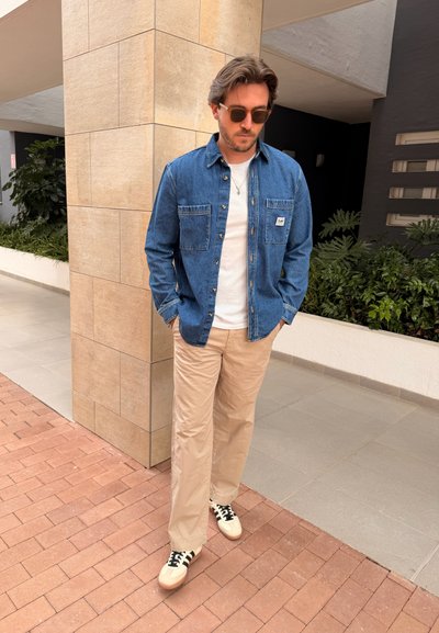 Hombre con gafas de sol que lleva una chaqueta vaquera, camisa blanca, pantalones beige y zapatillas blancas, de pie en un camino exterior con baldosas cerca de plantas.