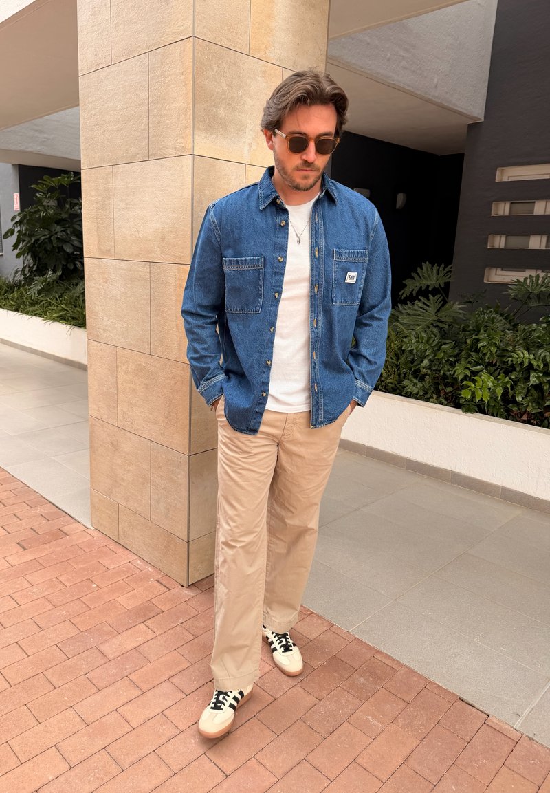 Homme avec des lunettes de soleil portant une veste en jean, une chemise blanche, un pantalon beige et des baskets blanches, debout sur un chemin extérieur carrelé près des plantes.