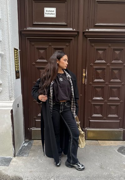 Abrigo largo negro sobre camiseta negra, camisa de cuadros y jeans negros con costuras blancas. Zapatos negro y gris. Bolso dorado. Fondo de puerta marrón.