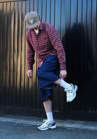 Camisa a cuadros roja y blanca, pantalones cortos de mezclilla oscuros y sueltos, calcetines blancos y zapatillas deportivas blancas. La gorra presenta un patrón de cuadros con un logo.
