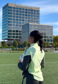 Osoba u svijetlozelenom i sivom sportskom odijelu drži nogometnu loptu, okrenuta prema modernom hotelu s staklenim prozorima na plavom nebu.