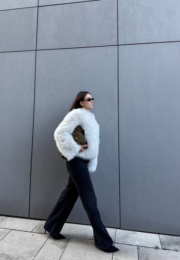Woman walking beside a gray tiled wall, wearing a white fluffy jacket, black wide-leg trousers, black heels, sunglasses, and holding a brown bag.