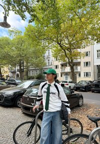 Chemise blanche à manches longues avec accents noirs et verts, cravate verte, jean bleu à jambes larges et casquette verte. Entouré de véhicules et d'arbres.