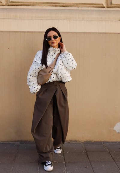 Mujer con chaqueta acolchada de lunares, pantalones anchos marrones, zapatillas deportivas, bolso bandolera beige y gafas de sol, de pie junto a una pared beige.