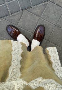 Mocassins en cuir marron portés avec des chaussettes blanches en dentelle et une jupe beige ornée d'une bordure en dentelle blanche, debout sur un trottoir carrelé gris.