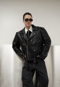 Veste de moto en cuir noir avec fermetures éclair argentées et épaulettes, portée avec une chemise blanche et une cravate noire. Pantalon en jean foncé porté.