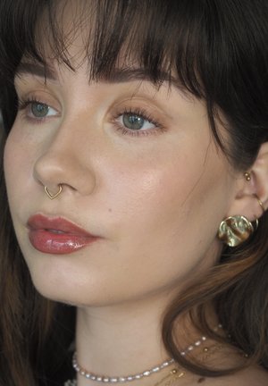 Gold heart-shaped nose ring, glossy lips, and gold earrings. Neutral skin tone, light eye makeup, fine hair with fringe, and layered necklaces.