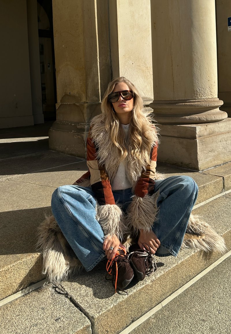 Long-sleeved, multi-colored coat with faux fur collar; gray sweater; light blue wide-leg jeans; dark shoes with orange accents. Sunlit setting.
