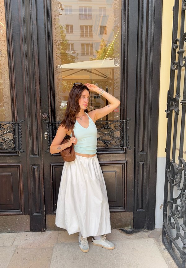 Joven mujer con falda blanca y top azul claro está junto a una puerta de madera oscura, sosteniendo unas gafas de sol sobre sus ojos, con bolso marrón al hombro.