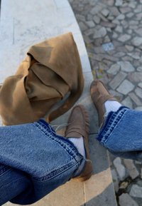 Hnedé semišové mokasíny s detailemi strapcov, v kombinácii s bielymi ponožkami a modrými rifľami. Na kamennom povrchu vedľa nich leží hnedá taška.